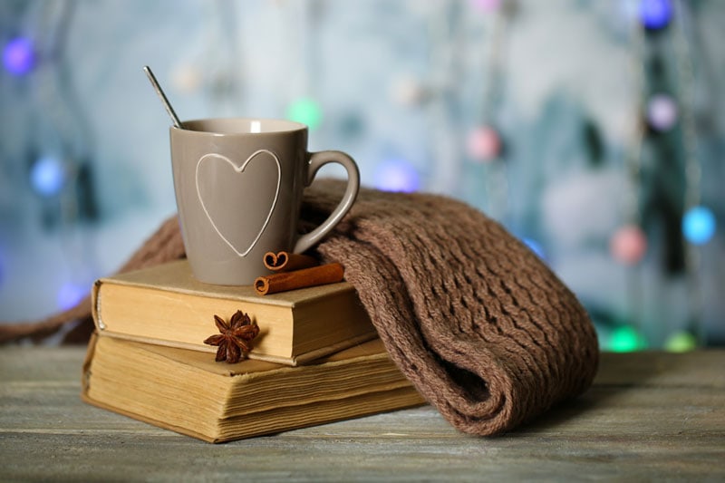 Tasse à cœur posée sur une pile de livres, accompagnée d'un plaid en laine, avec un arrière-plan lumineux et chaleureux.